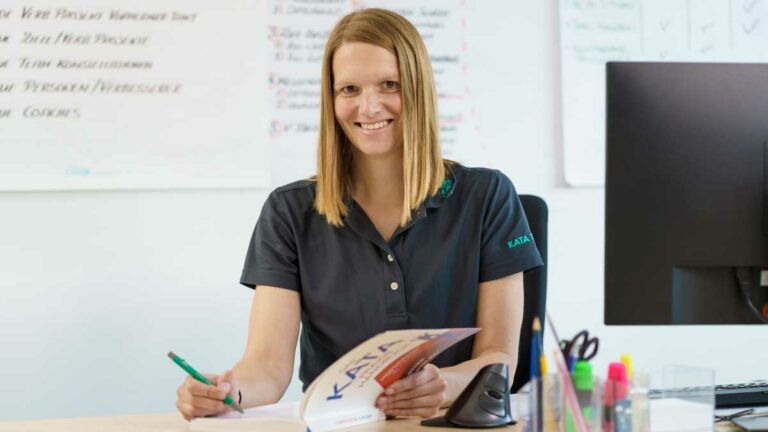 Frau Anne Heinig sitzt an ihrem Schreibtisch und hat ein Buch mit dem Titel "KATA" vor sich liegen. Rechts ist die Rückseite eines Bildschirms zu sehen. Auf dem Schreibtisch stehen in Utensilos Scheren und Stifte.