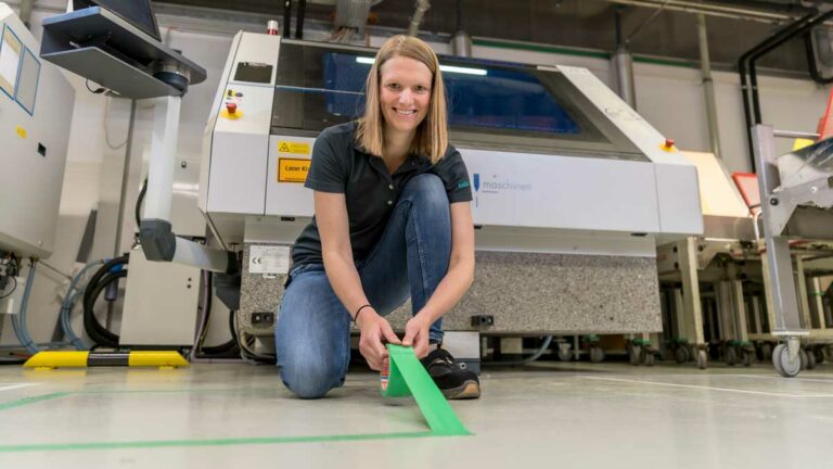 Ms. Anne Heinig squats in front of a machine and sticks green tape on the floor.