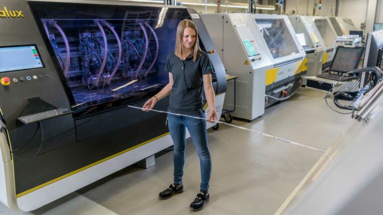 Anne Heinig steht zwischen Maschinen für die Leiterplattenproduktion und hält einen Zollstock in der Hand, mit dem sie den Abstand misst.